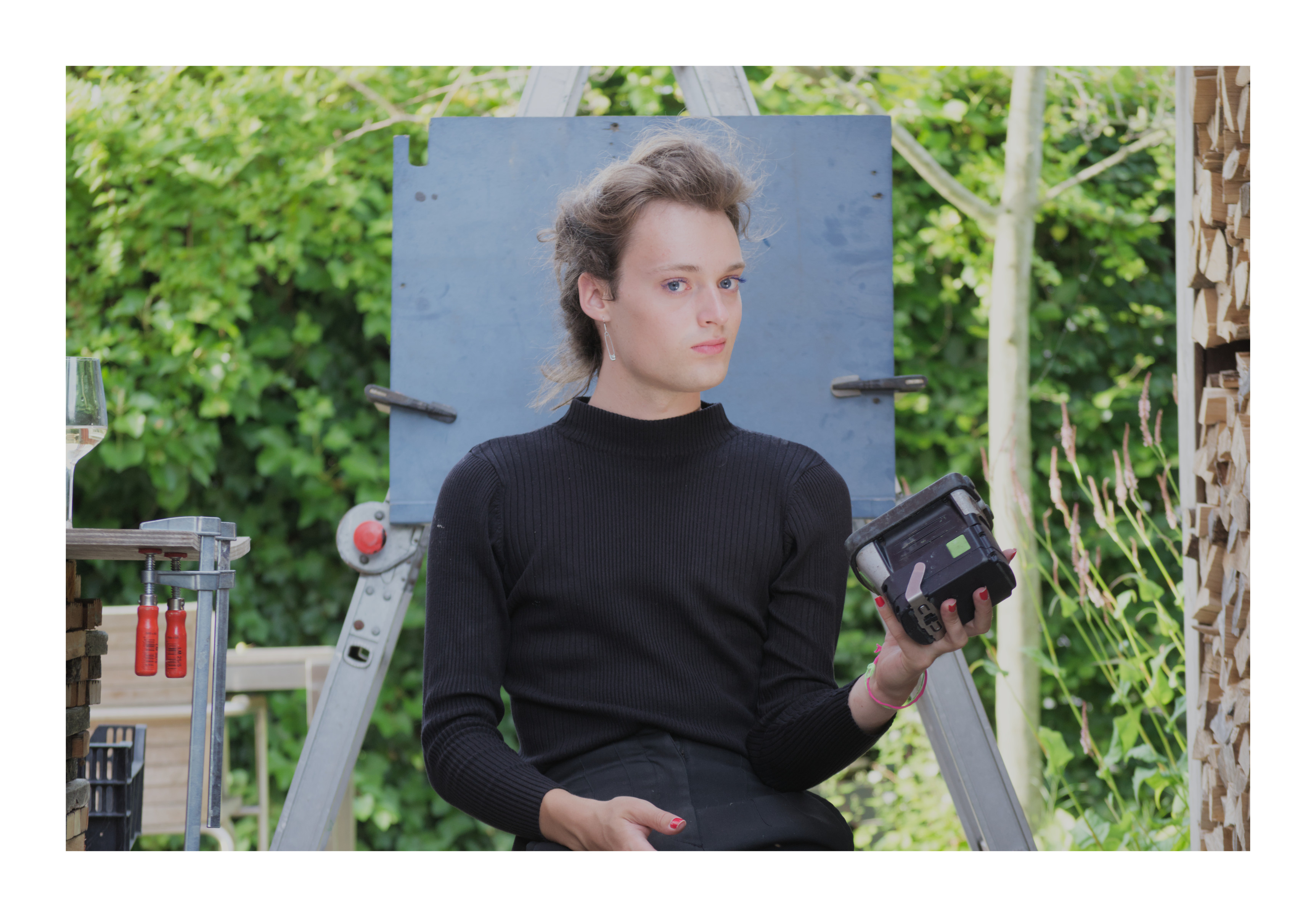 A portait of me, a white person with blue eyes and brown hair. I'm right in front of a pastel-blue painted sheet of wood suspended using clamps on a an aluminium ladder scaffold. I'm holding a workshop LED light in my left hand, shining onto my face.