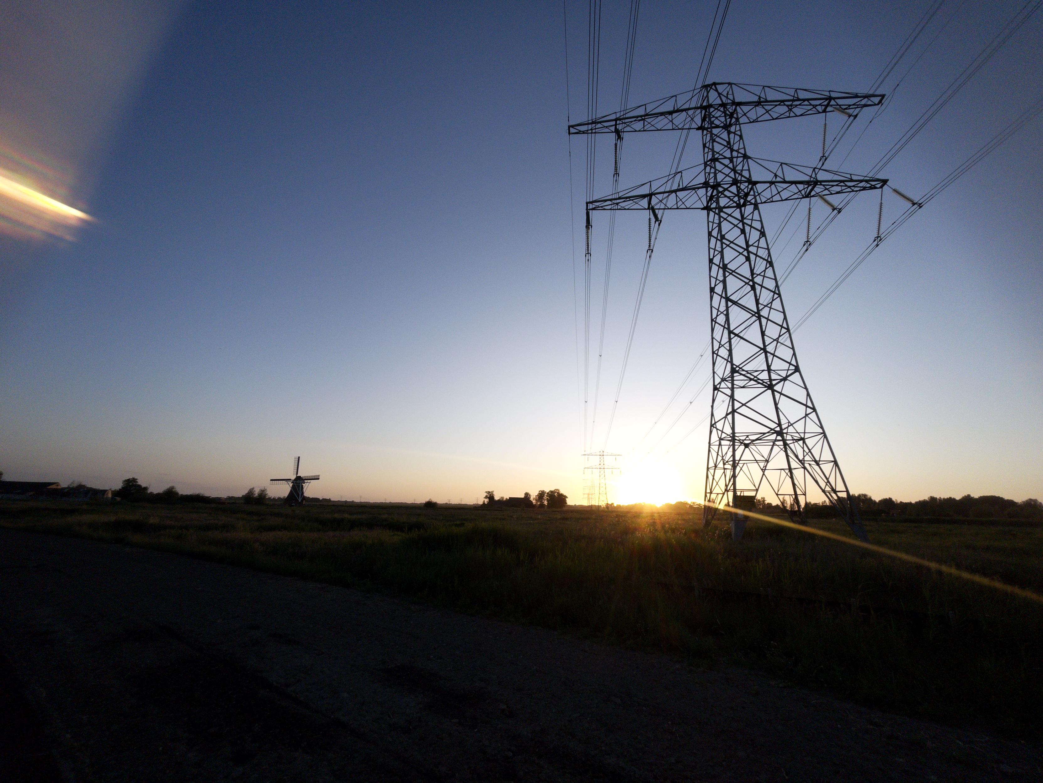 An old windmill and a powerline tower and overhead wires form silhouettes on a horizon with the sun setting. 