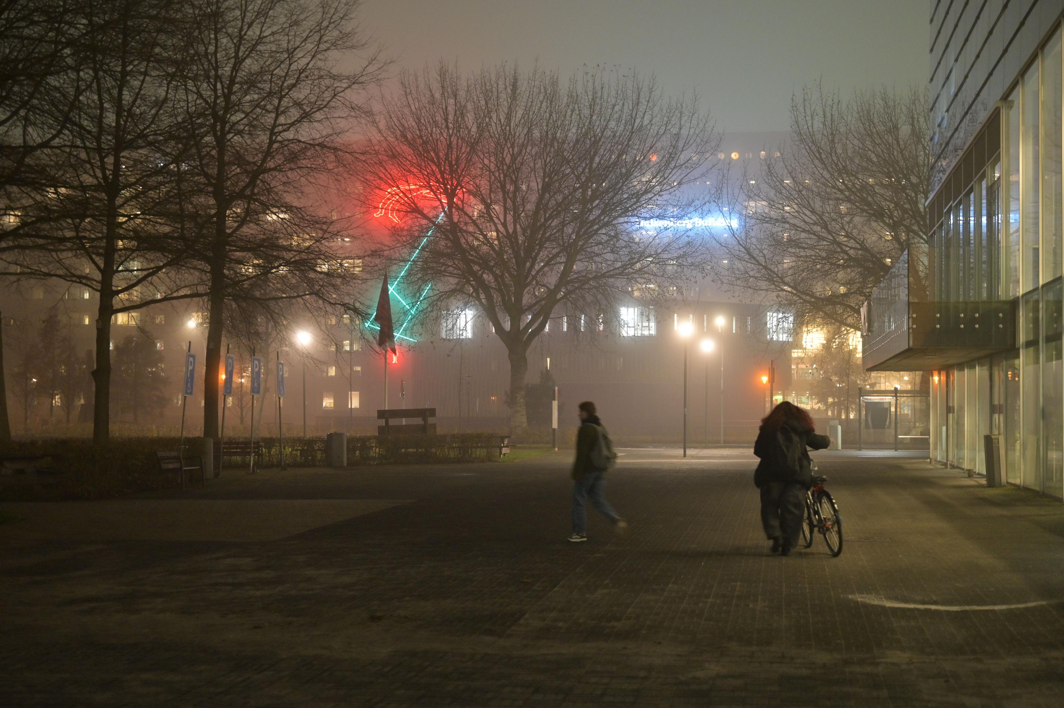 Standing next to the Bernoulliborg, a photo of the Duisenberg building with its neon light artwork shining beautifully through the fog, obstructed by the leafless trees. Two people walk past in the foreground.
