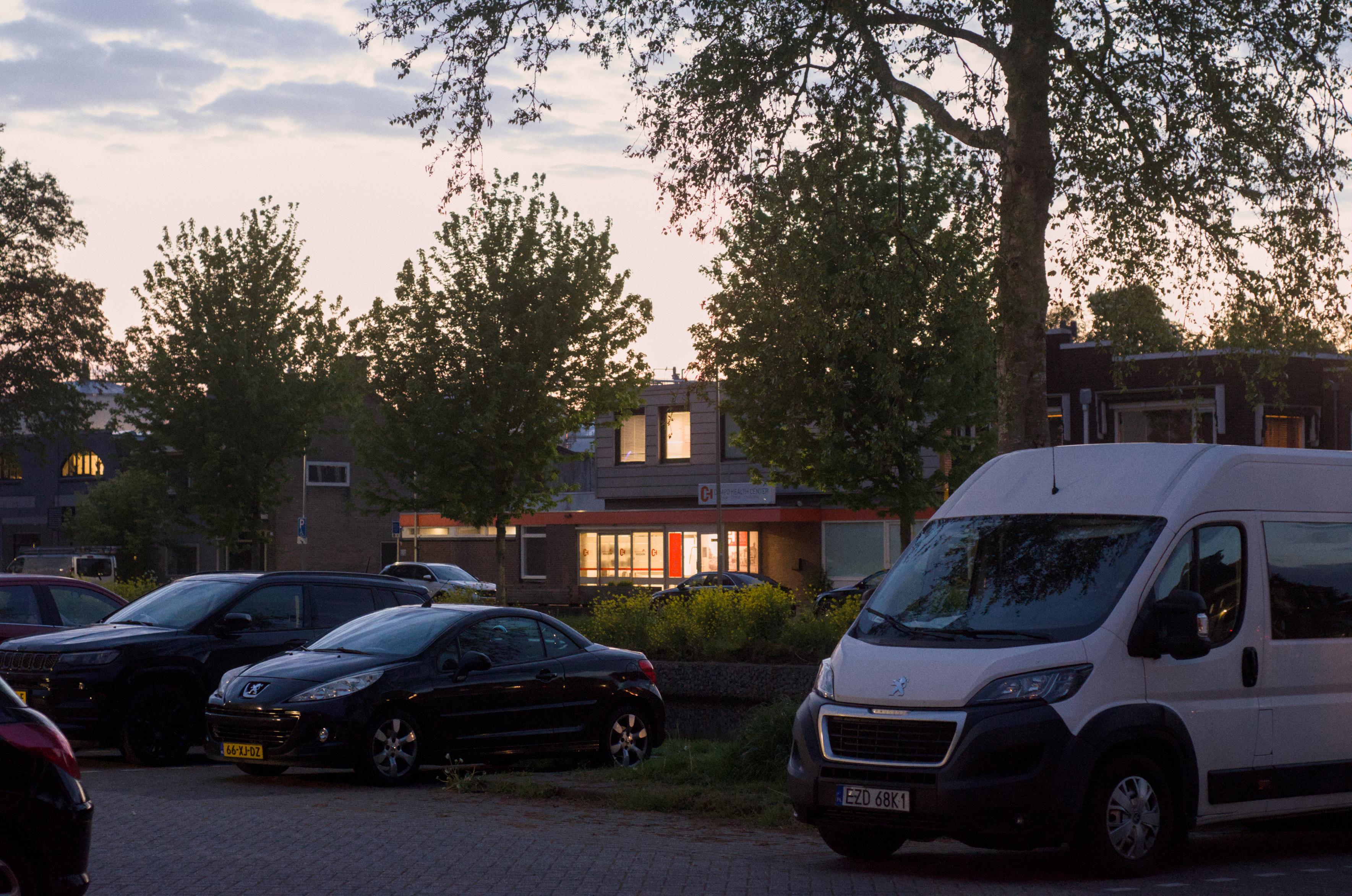 A warmly lit interior of a building is seen from the other side of a canal, standing among cars in a small parking lot.